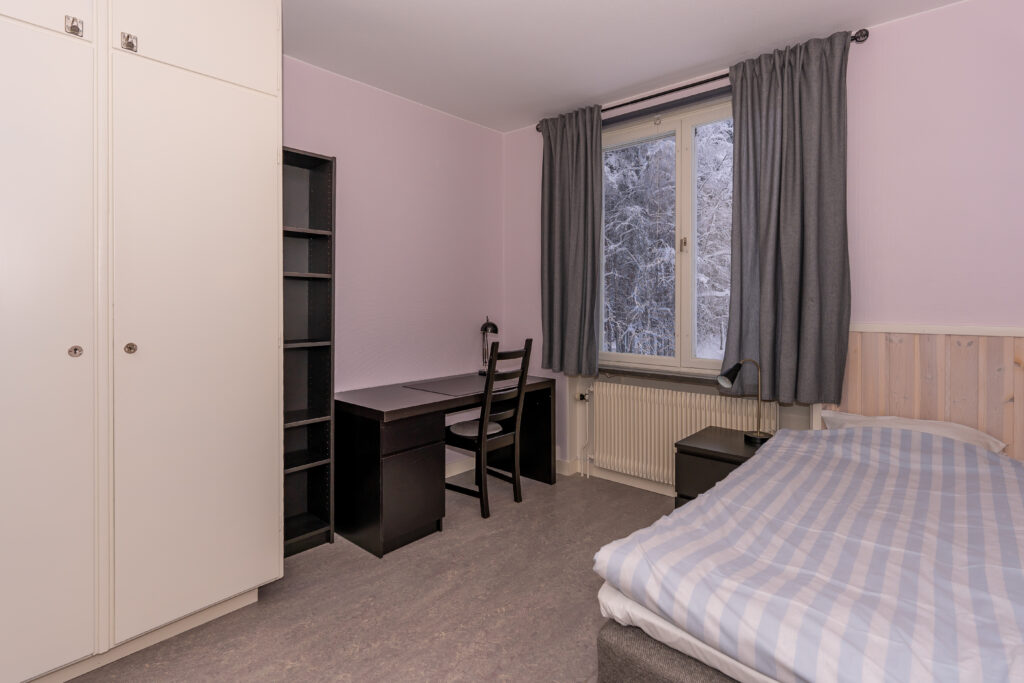 Rum med garderober, bokhylla, skrivbord med stol, säng och fönster.