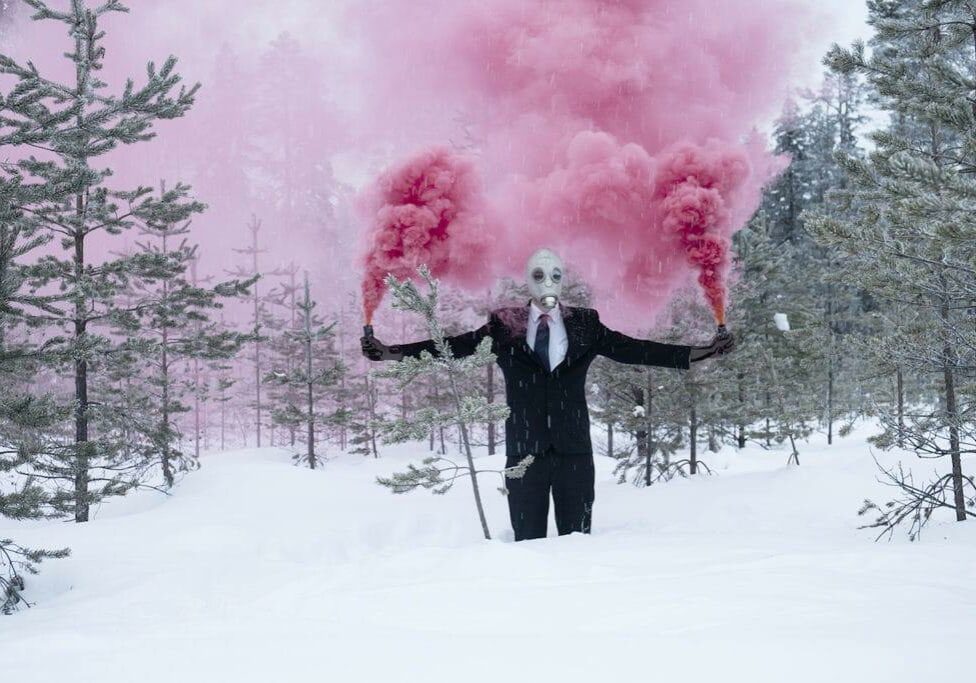 Person i gasmask och kostym håller två rosabolmande rökpatroner i händerna medan hen står till knäna i djup snö i tallskog.